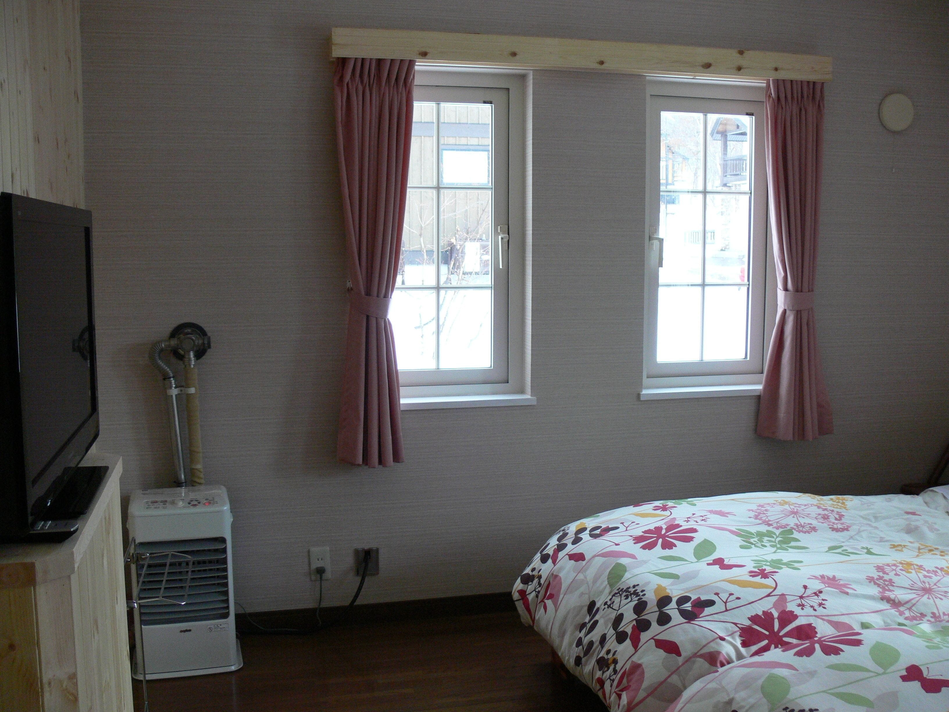 A bedroom with two windows, a bed with a floral bedspread, a heater, and a television on a wooden cabinet.