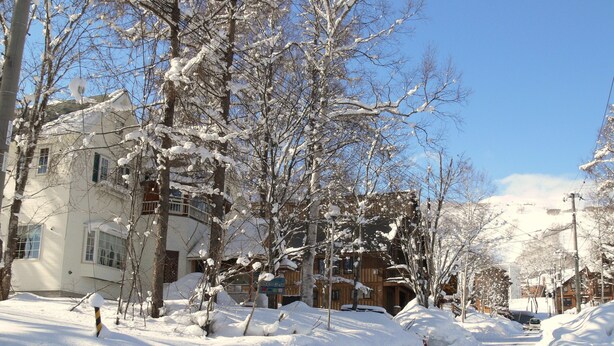 A snow-covered street with buildings, including a multi-story structure with balconies and a wooden cabin. - Pension Locomotion (Kutchan)