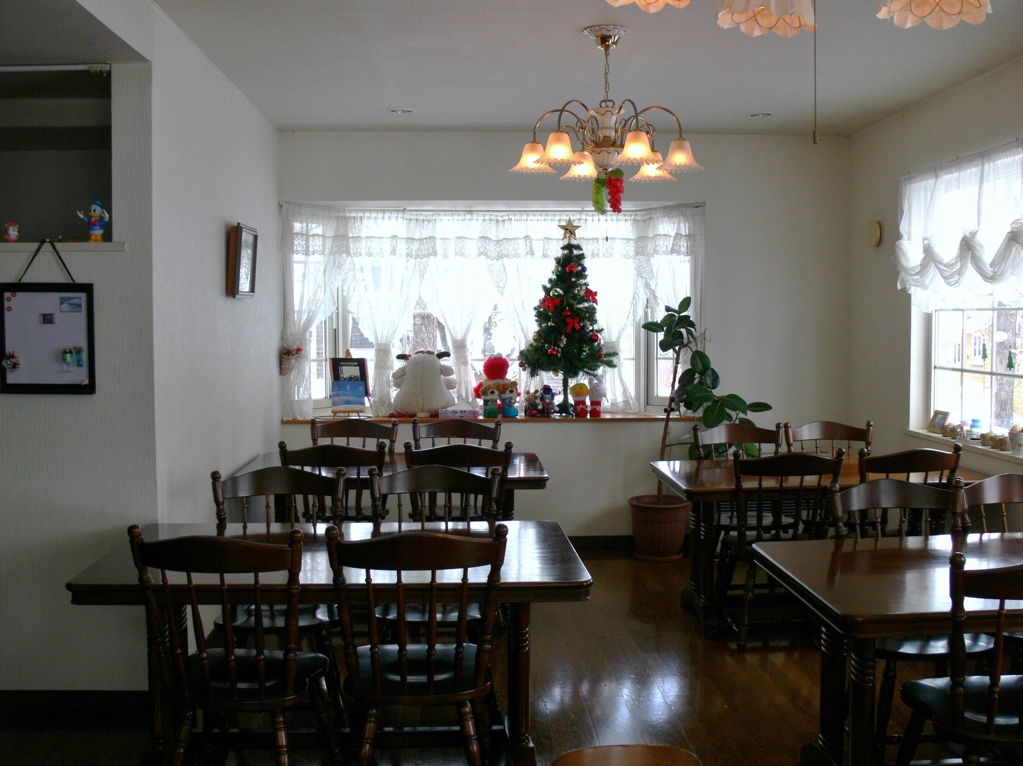 A dining area with wooden tables and chairs, a chandelier, a Christmas tree, and a window with lace curtains.