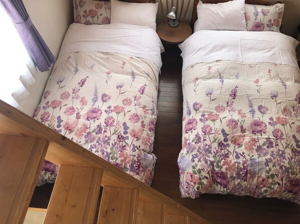 A bedroom with two beds, each with floral bedding and white pillows, a bedside table with a lamp, and a wooden staircase in the foreground. - Pension Locomotion (Kutchan)