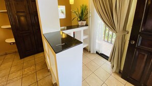 A kitchen with a black countertop, white cabinets, a potted plant, and a view through a window with curtains. - Hotel Puerto Libre (Puerto Barrios)