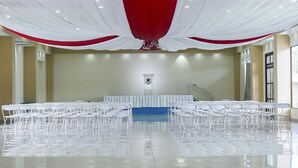 A large, well-lit hall with white chairs arranged in rows, a stage with a blue backdrop, and a red and white draped ceiling. - Hotel Puerto Libre (Puerto Barrios)