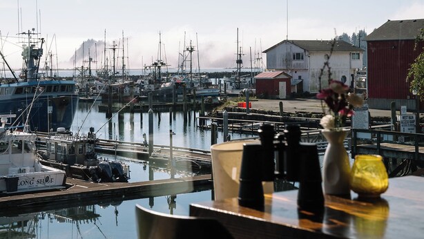 A view from inside a building looking out to a marina with boats docked, a table with chairs, and a vase with flowers. - Salt Hotel & Pub (Ilwaco)