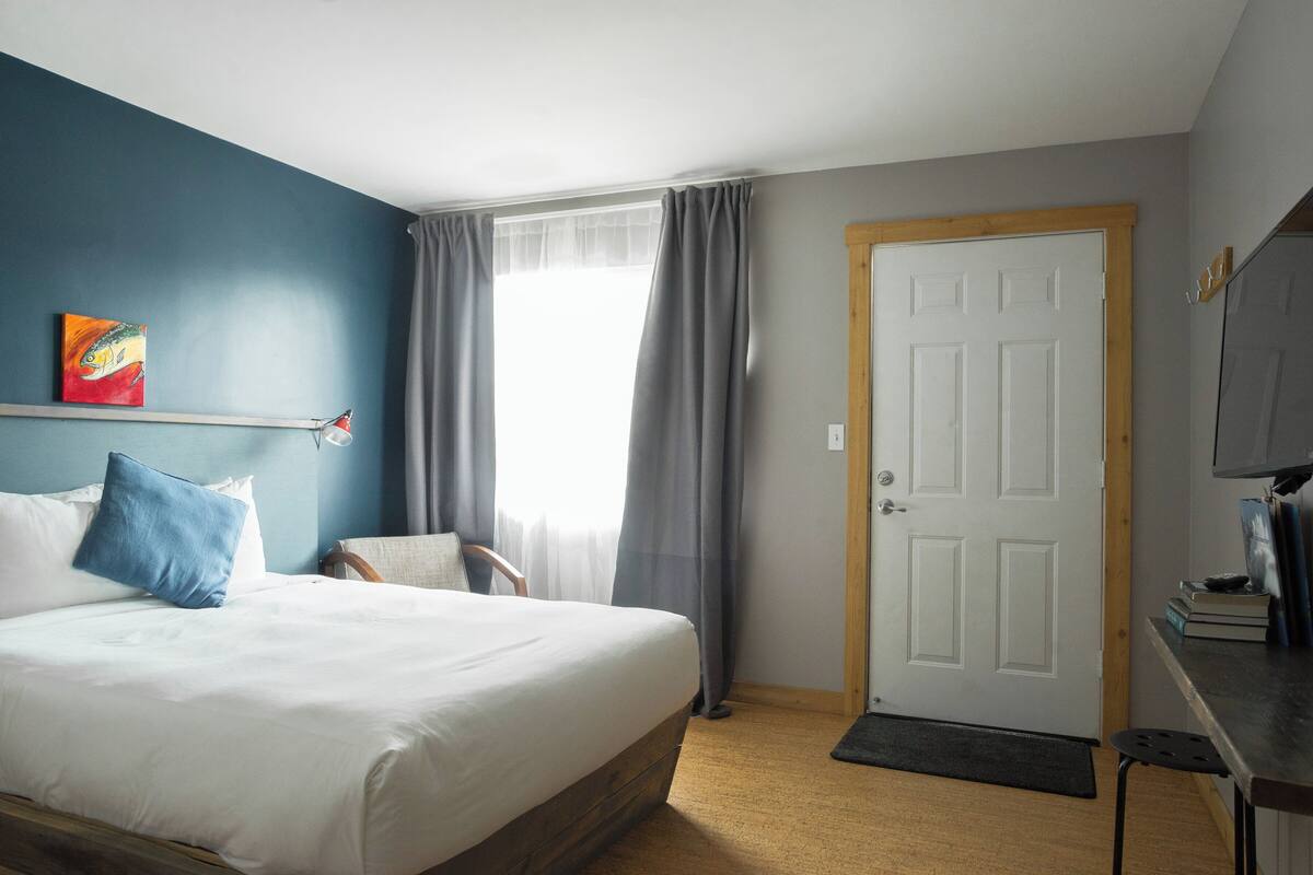 Hotellihuone, jossa on sänky, tuoli, työpöytä, ovi ja ikkuna, jossa on verhot.