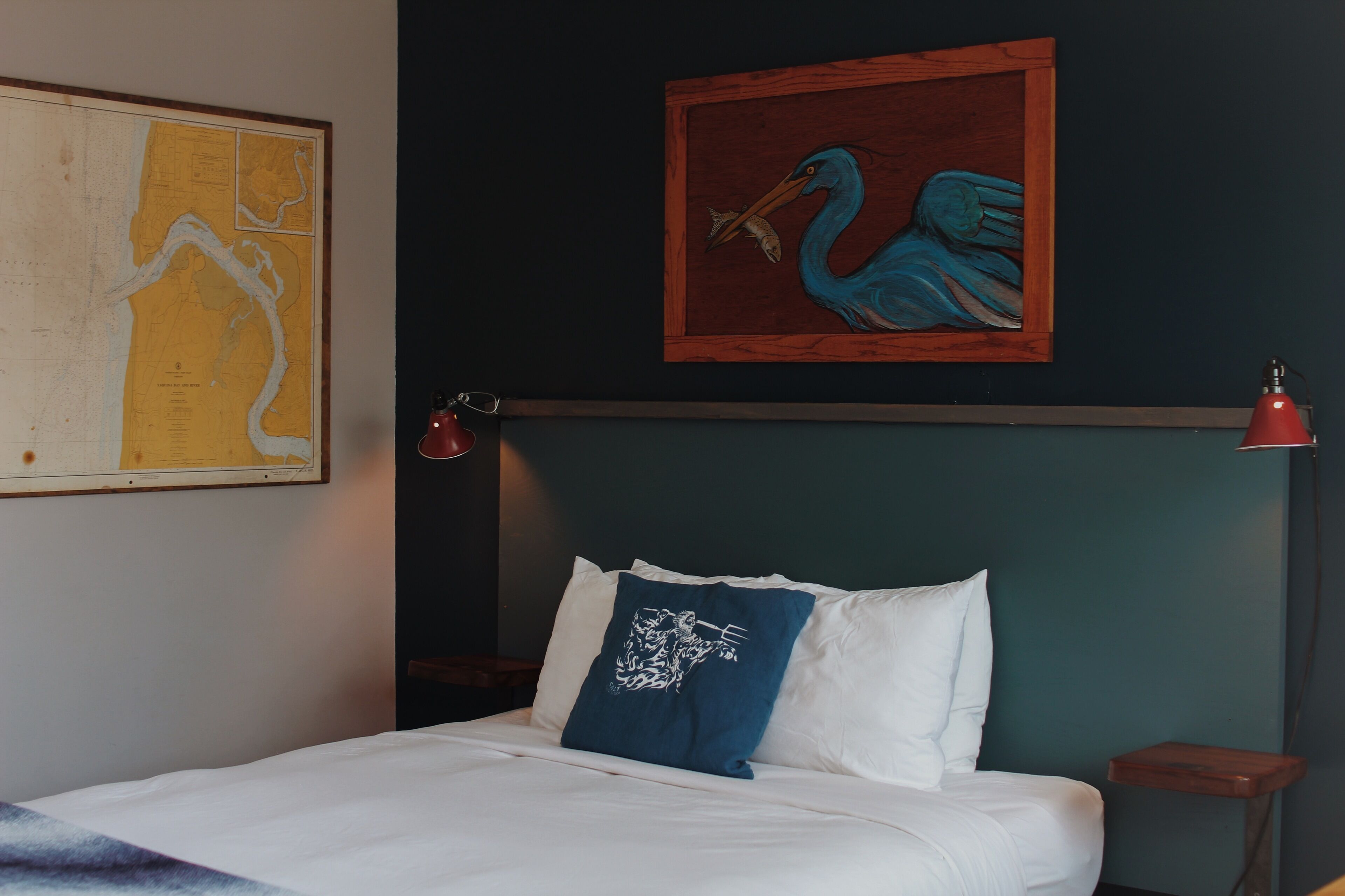 Una cama con ropa de cama blanca, una almohada azul con diseño blanco, una lámpara roja sobre una mesita de noche y un cuadro enmarcado de un pájaro azul en la pared.