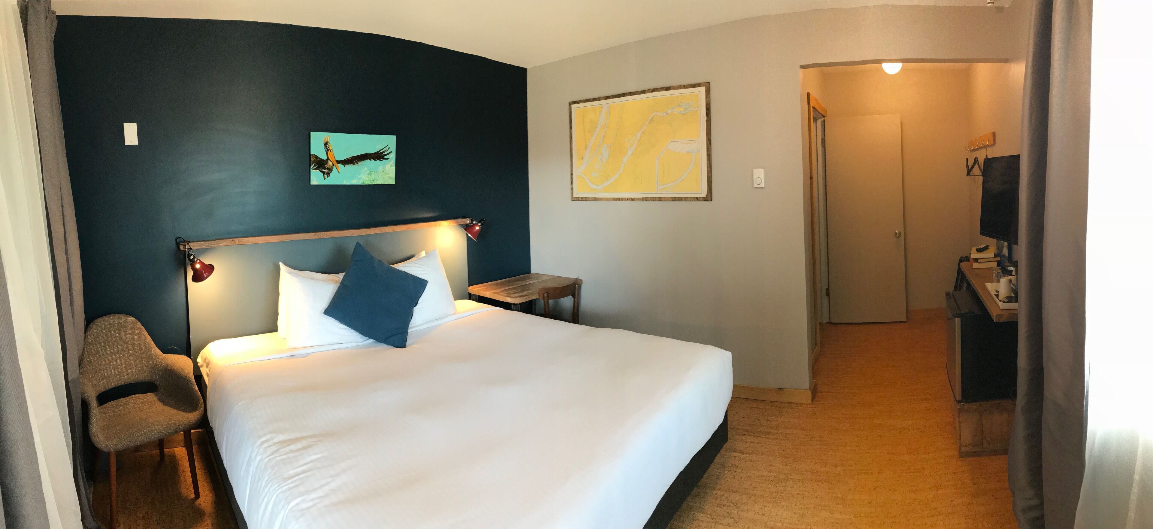 Et hotelværelse med en stor seng, et skrivebord med en computer, en stol, en skrivebordslampe og kunst på væggene.
