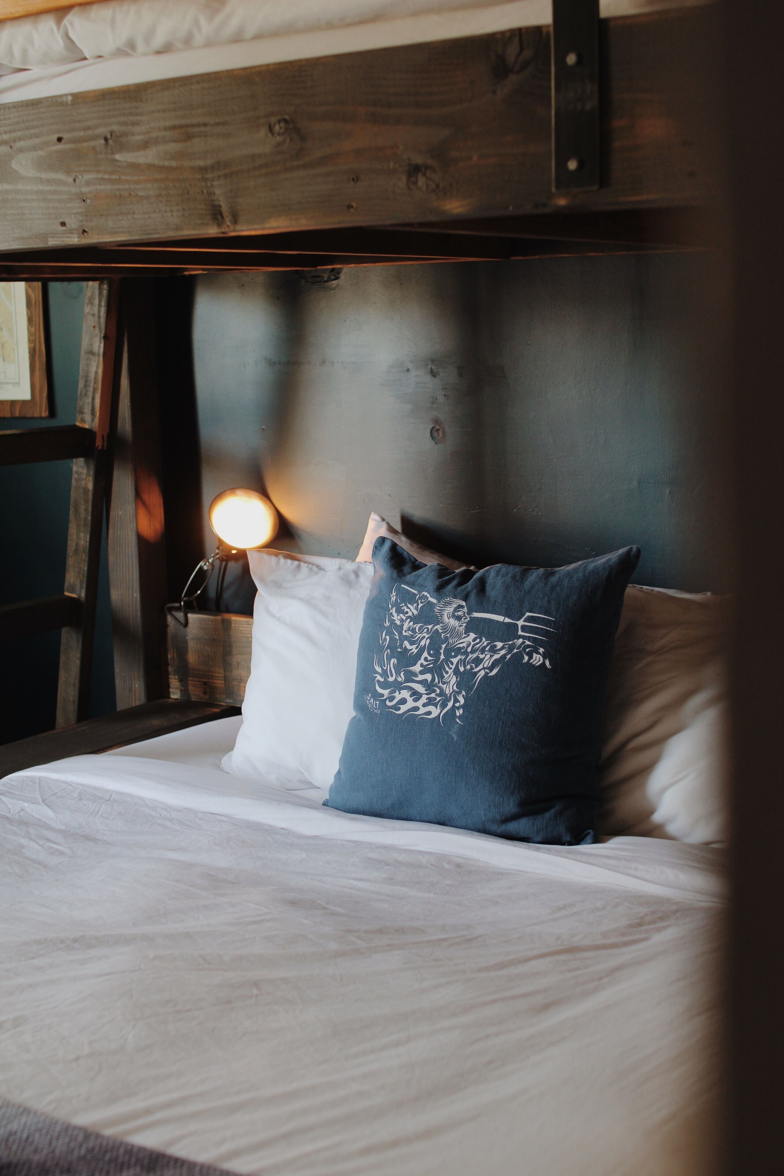 Una cama con ropa de cama blanca y un almohadón azul con diseño blanco, cabecera de madera y una lámpara fijada a la pared.
