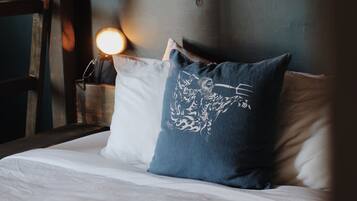 Una cama con ropa de cama blanca y un almohadón azul con diseño blanco, cabecera de madera y una lámpara fijada a la pared.