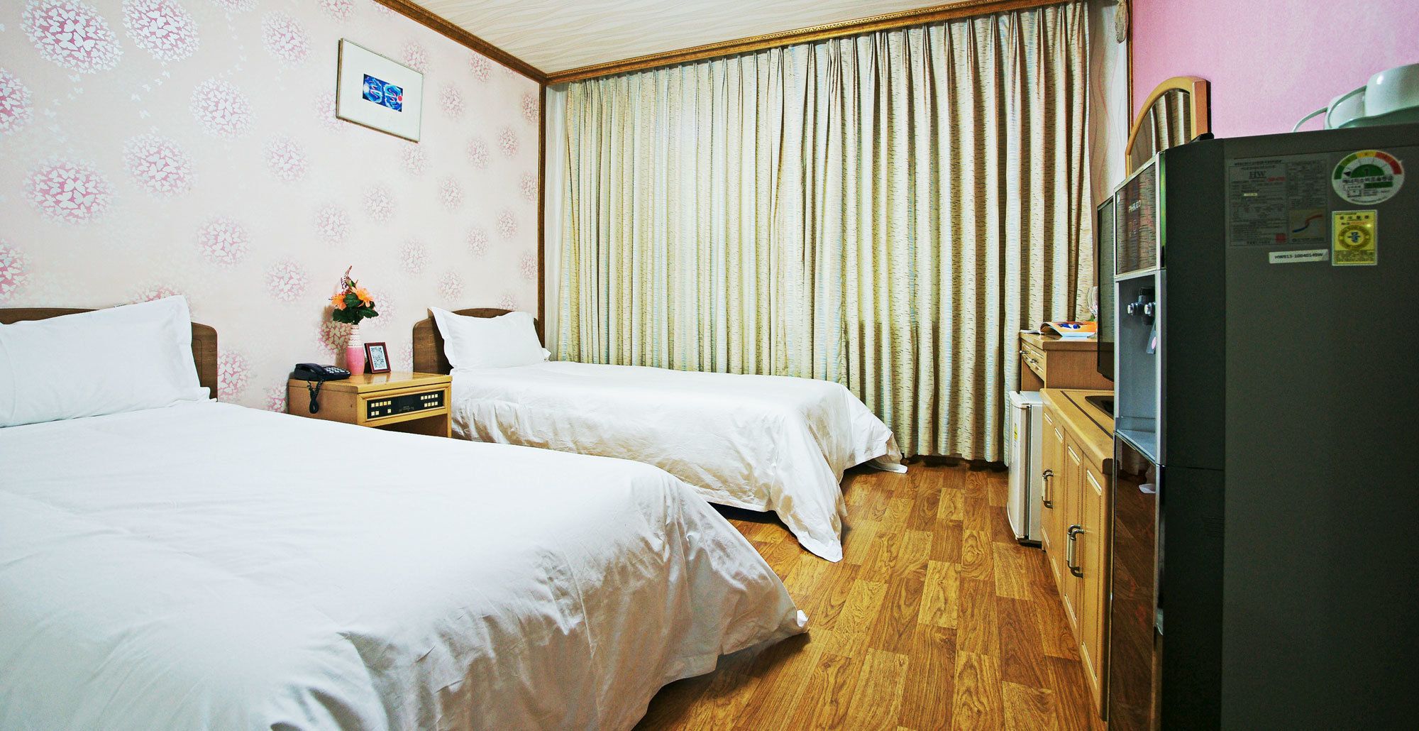 A hotel room with two beds, a wooden dresser, a refrigerator, and a window with curtains.