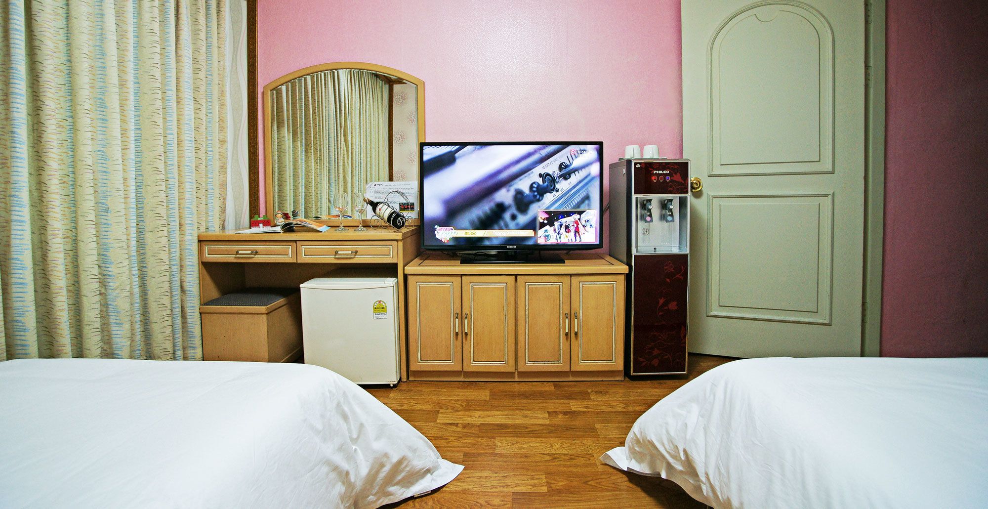Ein Hotelzimmer mit zwei Betten, einem Schreibtisch mit Computer, einem Kühlschrank, einer Minibar und einem Fernseher.