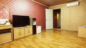 A room with a wooden floor, a television on a cabinet, a refrigerator, and a door leading to another room. - Emerald Hotel (Jeju City)