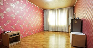 Una habitación con piso de madera, teléfono, refrigerador y ventana con cortinas.