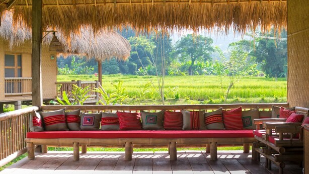 A wooden deck with a thatched roof, a bench with red cushions, and a view of a lush green field. - Lisu Lodge (Mae Taeng)