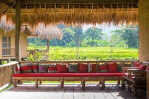 Una terraza de madera con techo de paja, un banco con cojines rojos y vistas a un campo verde exuberante.