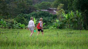 Two people walking in a grassy field with dense vegetation and a building in the background.