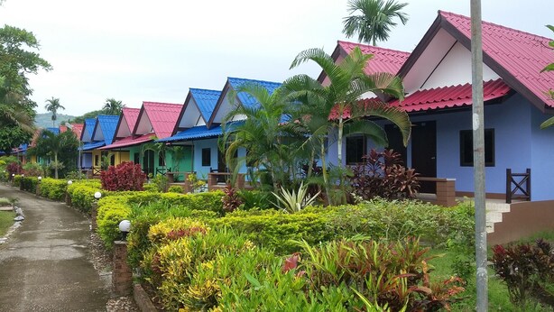 A row of colorful cottages with red, blue, and pink roofs, surrounded by lush greenery and a paved path. - Ruenview Resort (Mae Chan)