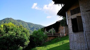 A rustic wooden house with a thatched roof, a balcony, and a view of a forested mountain.