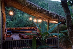 Eine Holzterrasse im Freien mit Sitzgelegenheiten und hängenden Lichtern, umgeben von üppigem Grün.