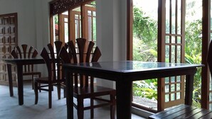A dining area with a glass-top table and wooden chairs, large windows with a view of greenery, and a wooden door with a lattice design. - Srimali's Residence (Unawatuna)