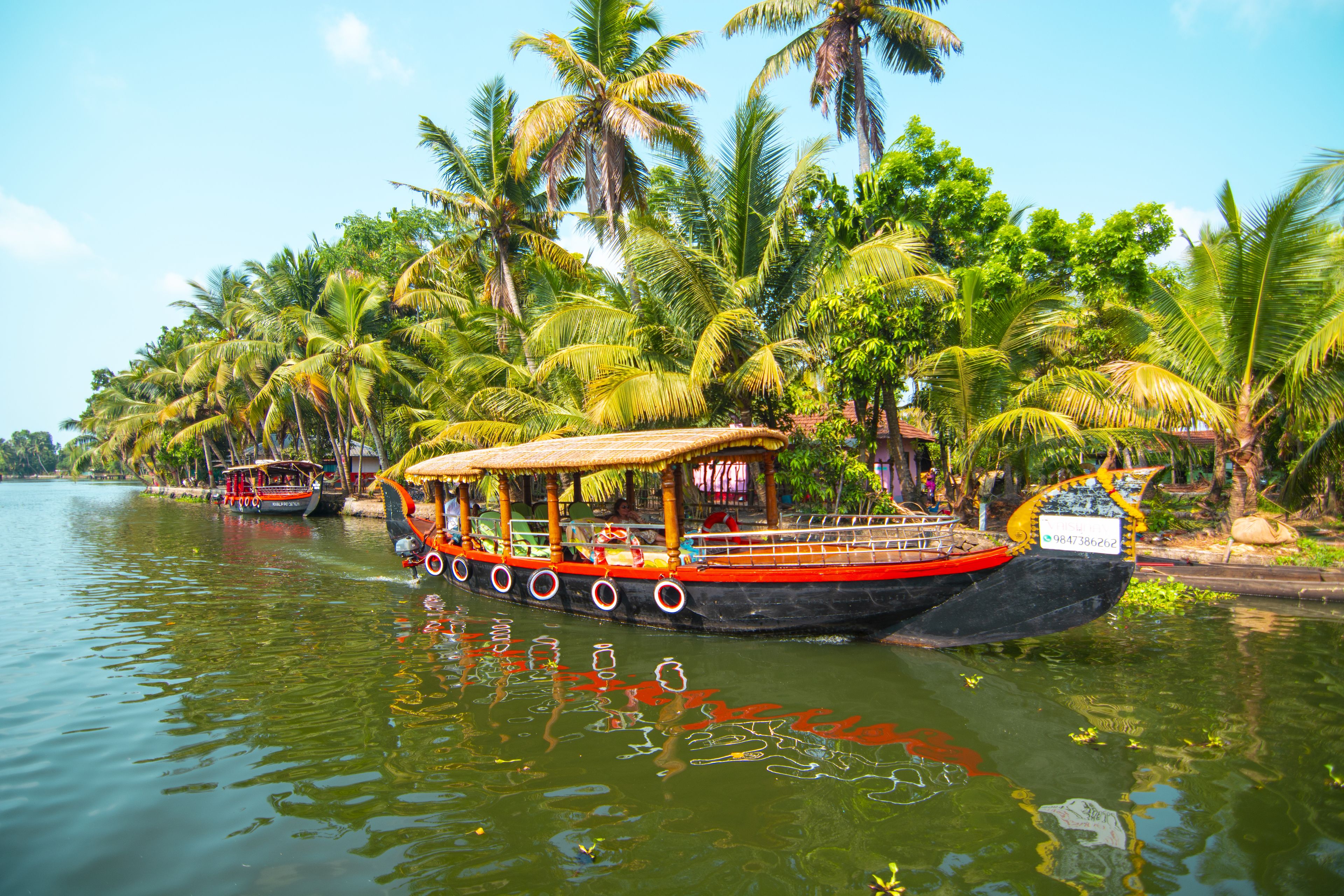 A boat with a thatched roof and a black hull, docked at a calm river surrounded by palm trees.