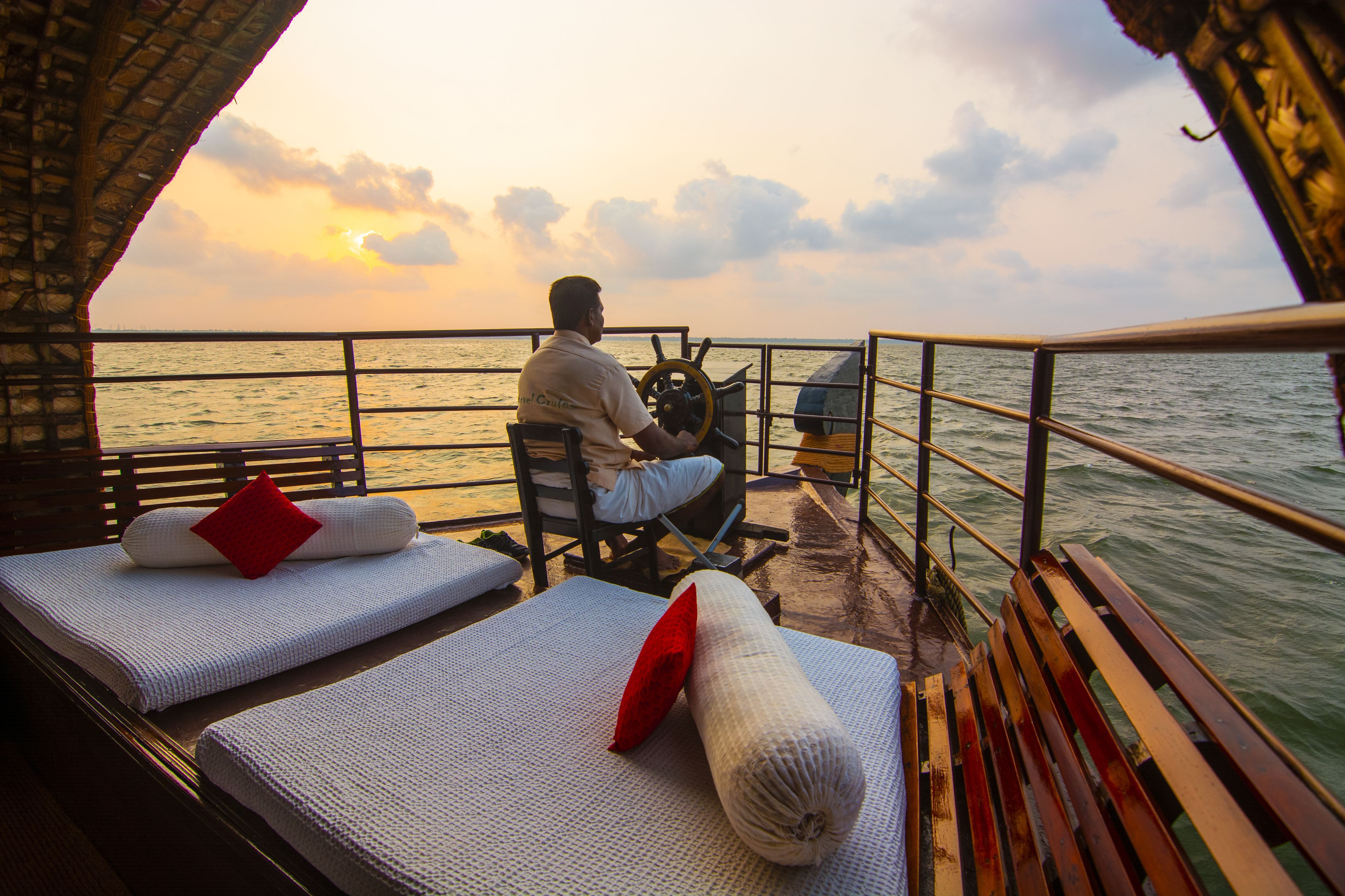 A boat deck with two reclining chairs, a steering wheel, and a view of the sunset over the water.