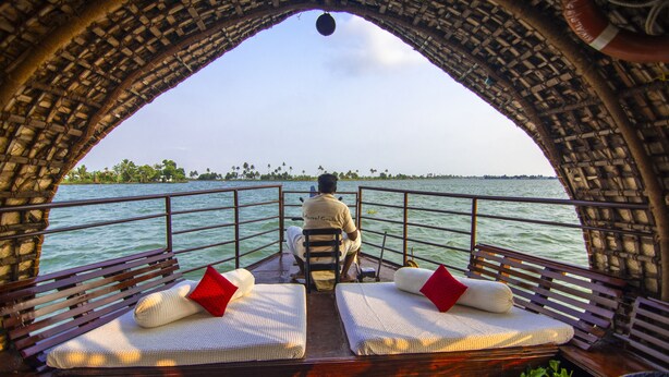 A boat with two cushioned seating areas, each with a red pillow, and a person seated at the back. - Marvel Cruise (Kuttanad Taluk)