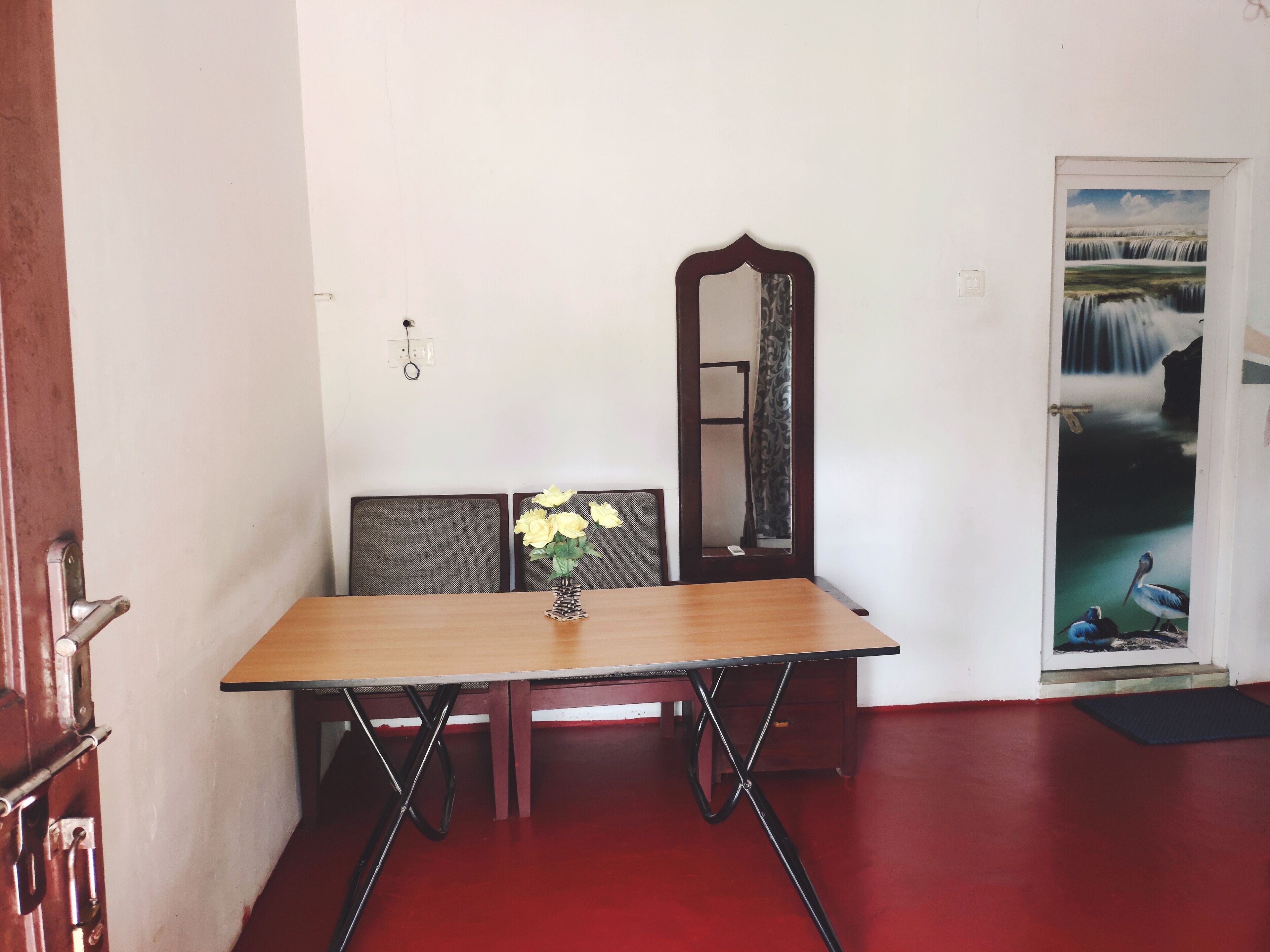 A dining area with a wooden table, two chairs, a vase with flowers, a mirror, and a door with a waterfall painting.
