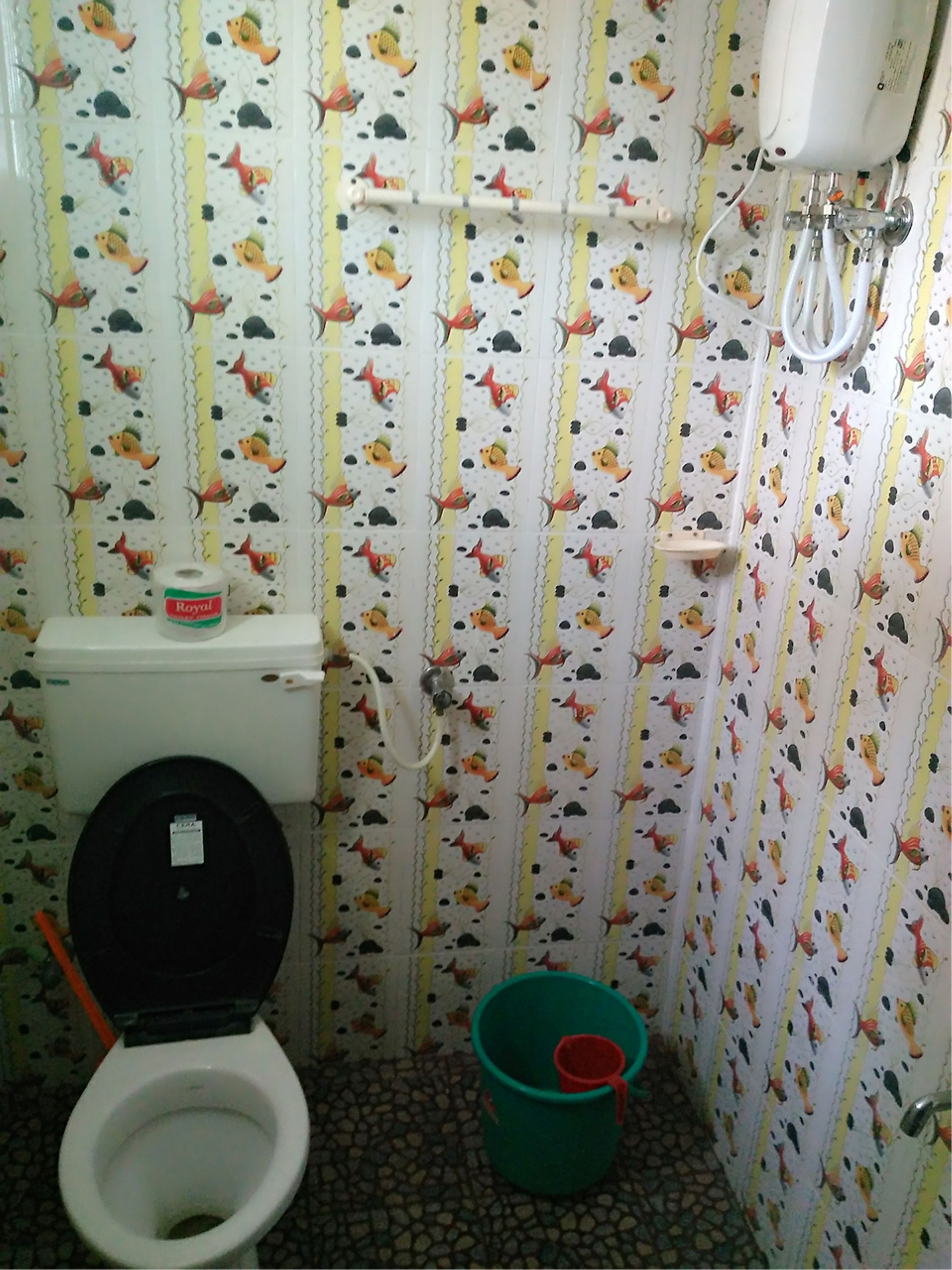 A small bathroom with a toilet, a green bucket, and a patterned wallpaper.
