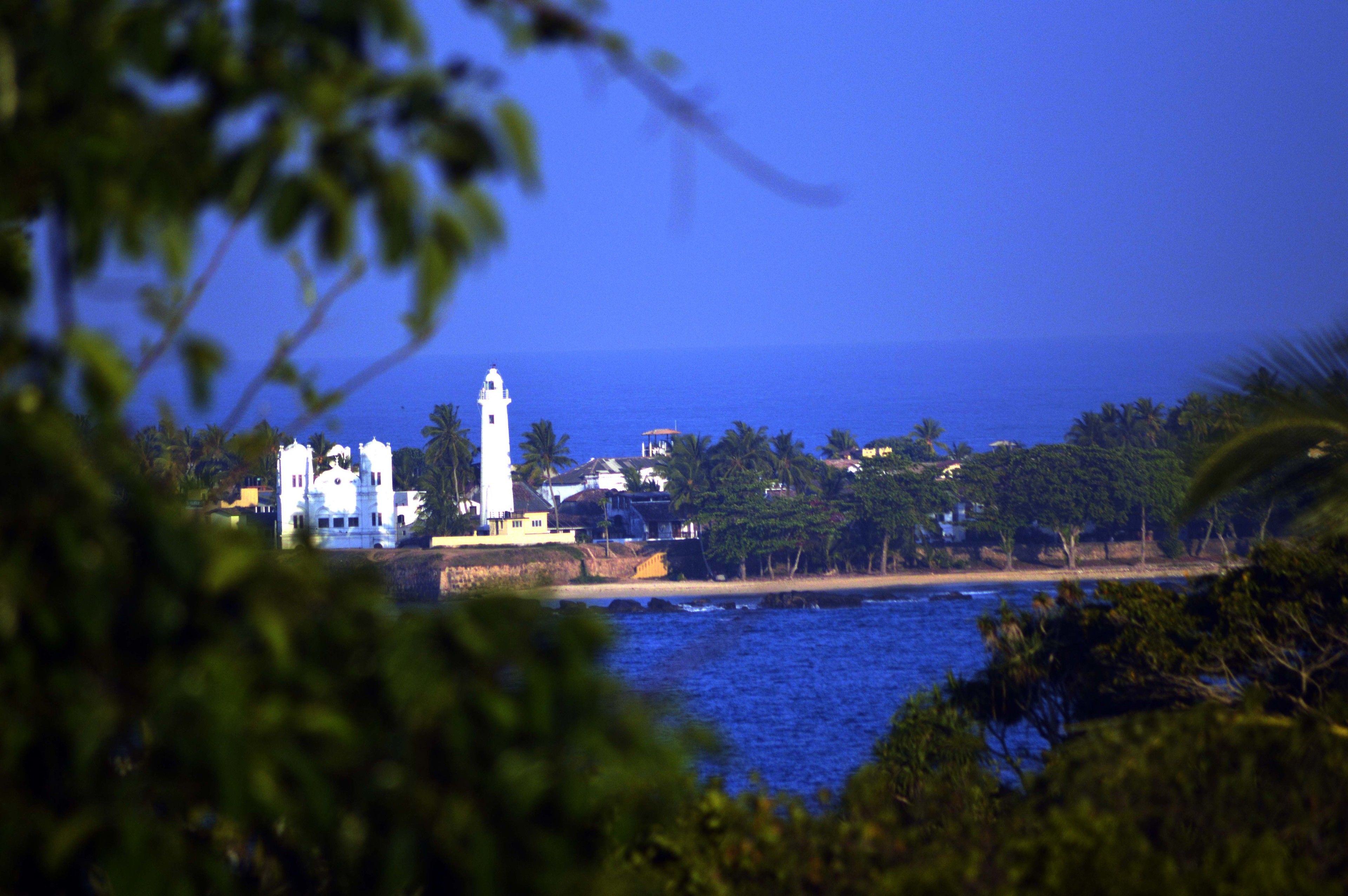 Vista costeira com um farol branco imponente, uma praia e edifícios residenciais.