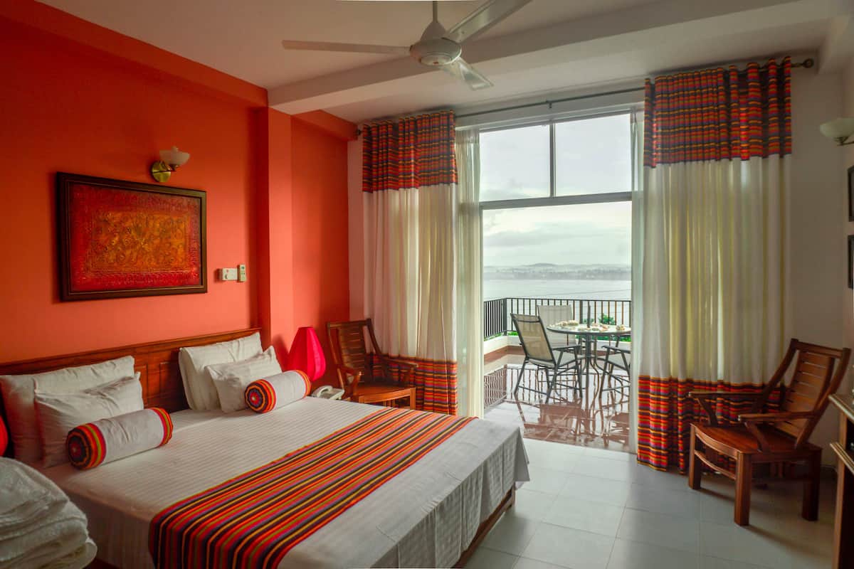 Habitación de hotel con una cama grande, ventilador de techo, balcón con mesa y sillas, y vista al mar.