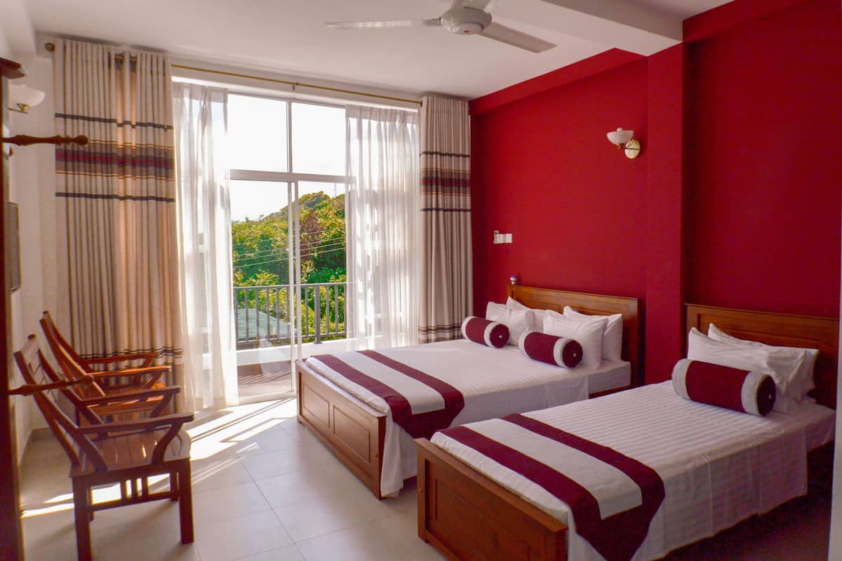 Habitación de hotel con dos camas, un balcón con cortinas, sillas de madera y una pared roja.