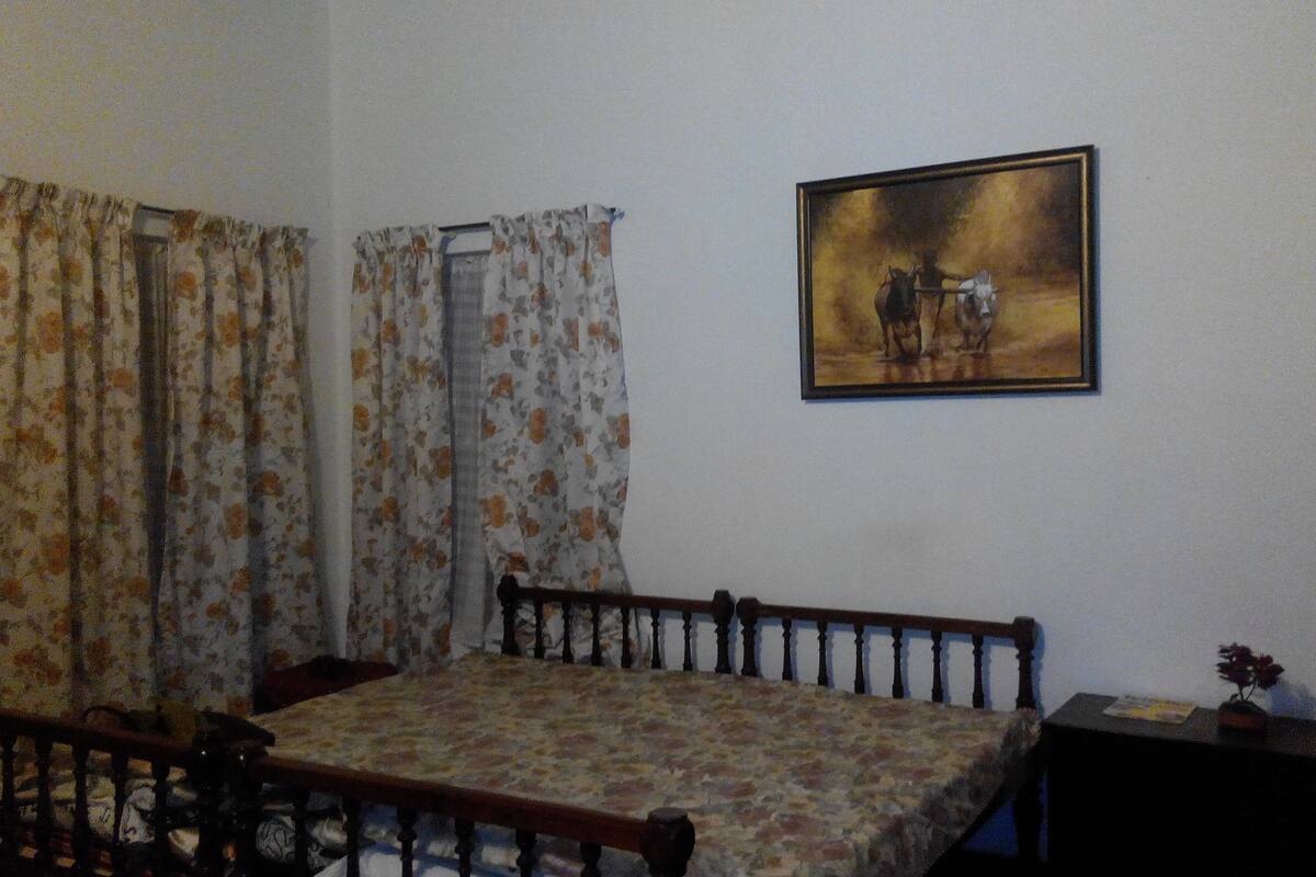 A room with a bed, a small table, a chair, a painting on the wall, and curtains.