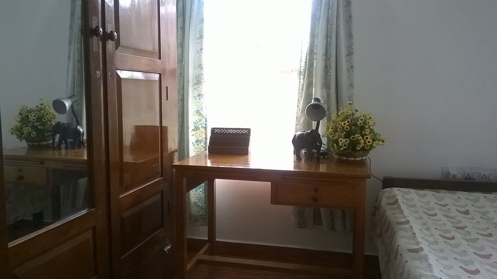 Een kamer met een houten bureau, een stoel, een bed met beddengoed met een patroon, een raam met gordijnen en een kleine plant op het bureau.