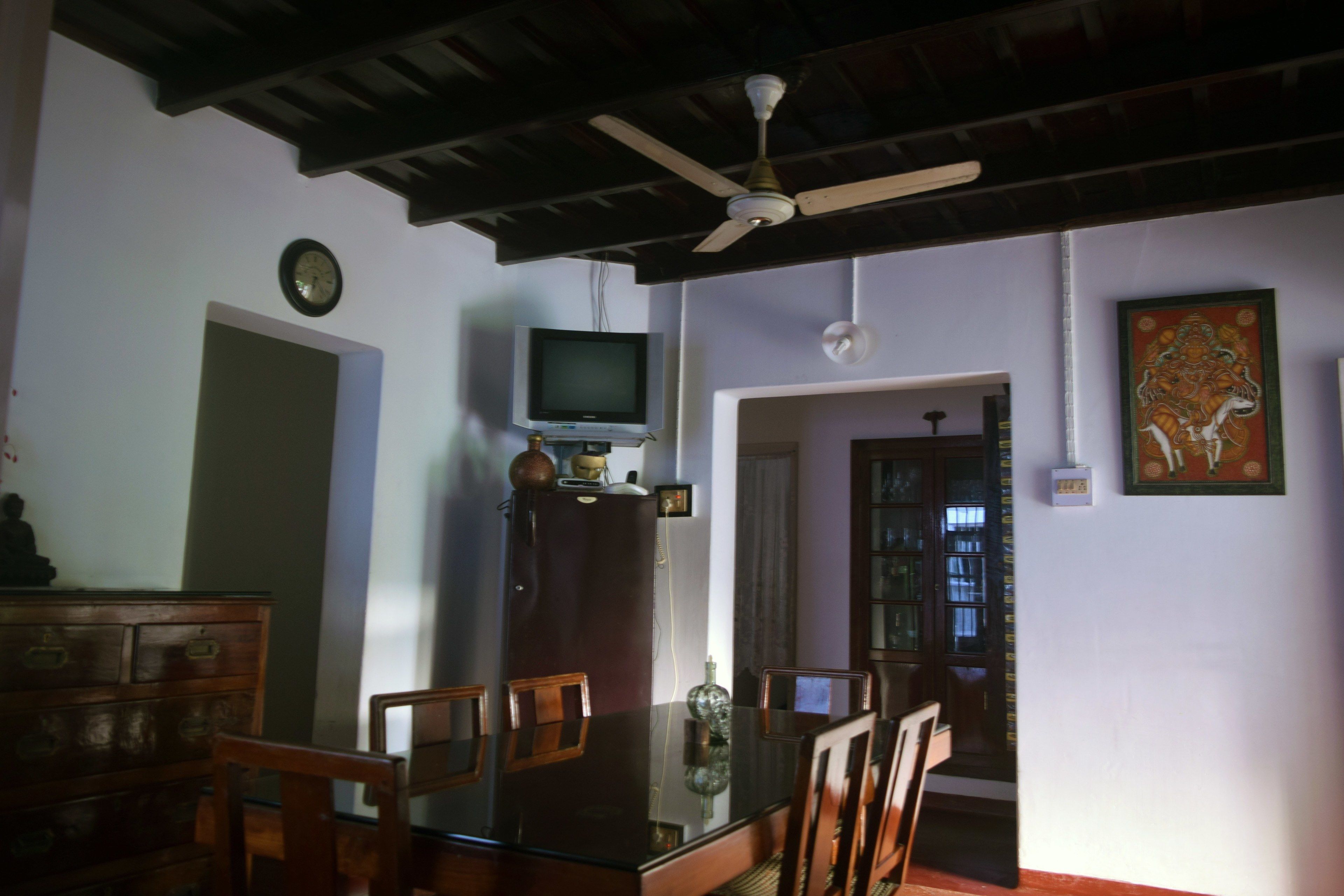 A dining area with a glass table, wooden chairs, a ceiling fan, a television on a cabinet, and a bookshelf.