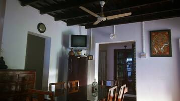 A dining area with a glass table, wooden chairs, a ceiling fan, a television on a cabinet, and a bookshelf.