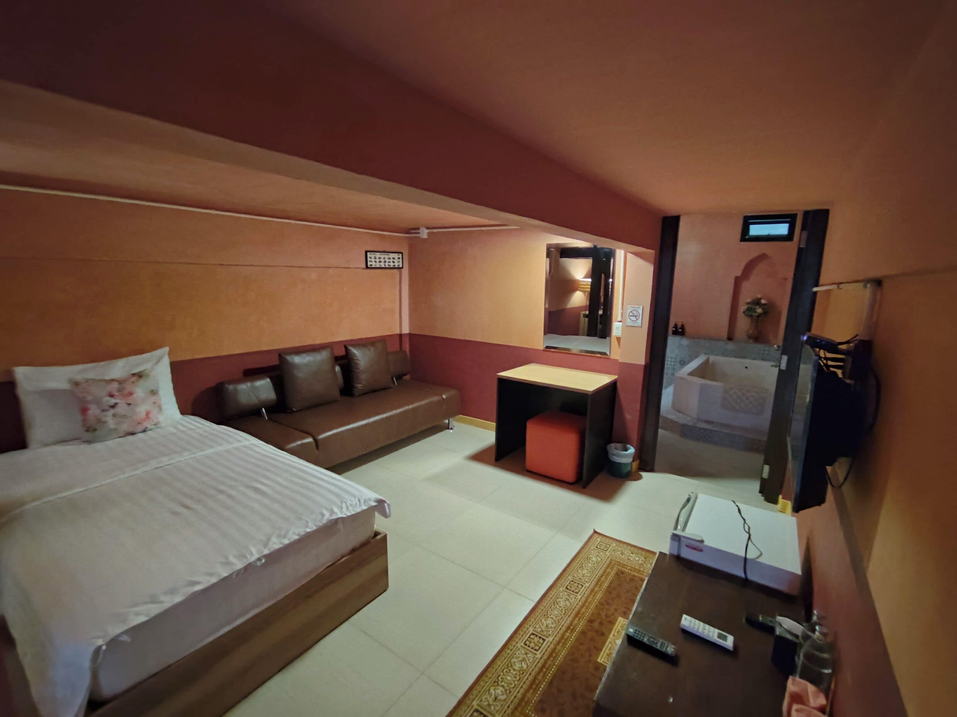 A hotel room with a bed, a sofa, a small table, a mirror, and a bathroom visible through an open door.