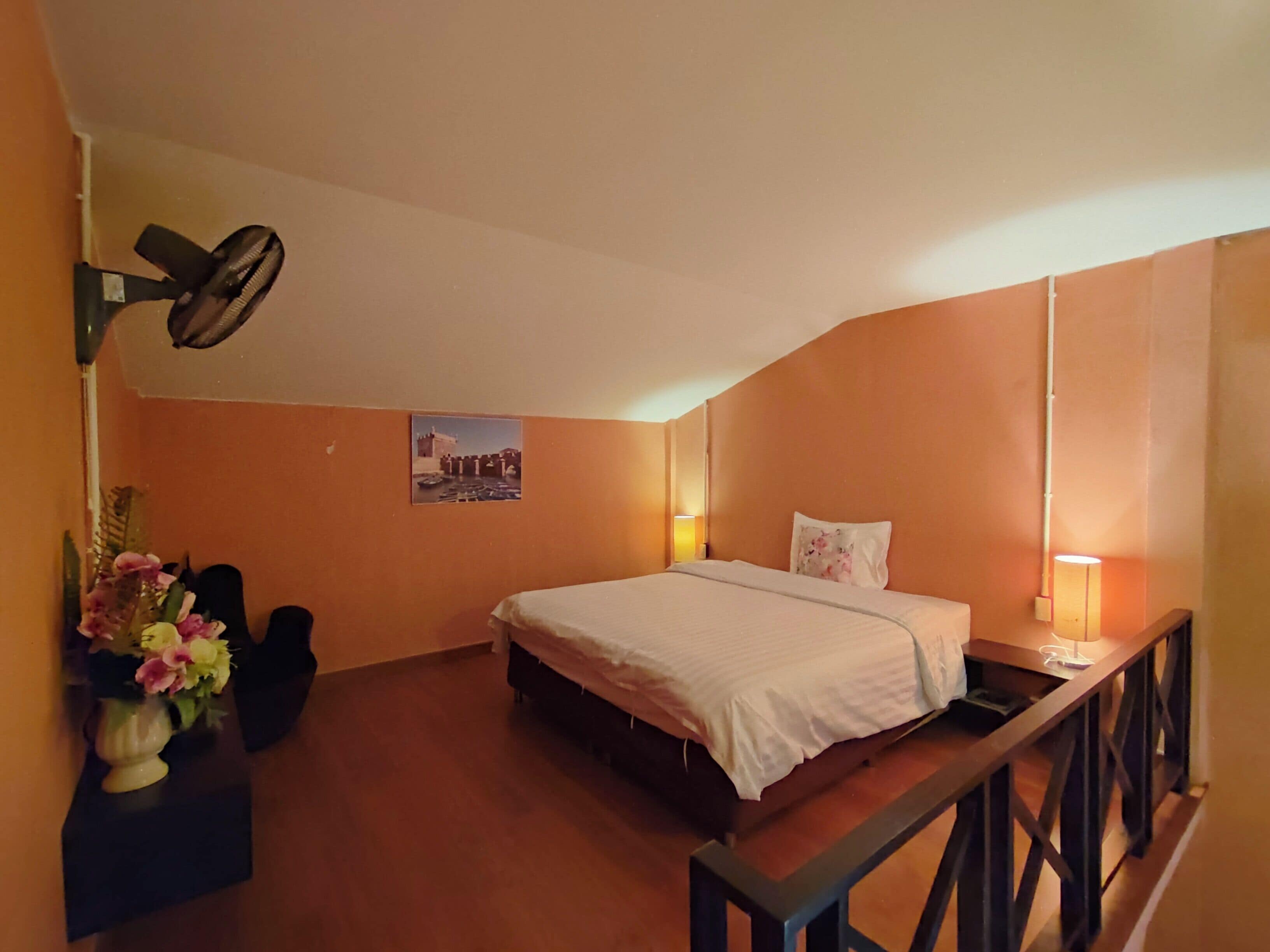 A bedroom with a bed, a desk with a lamp, a picture on the wall, and a ceiling fan.