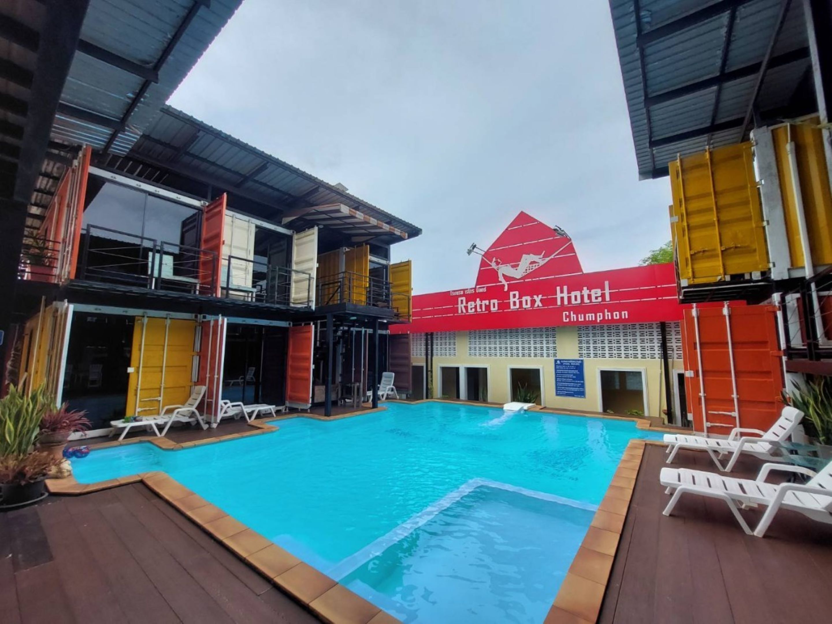 Ein Poolbereich mit Liegestühlen, einem Schild des Retro Box Hotels und einem Gebäude mit bunten Versandcontainern.
