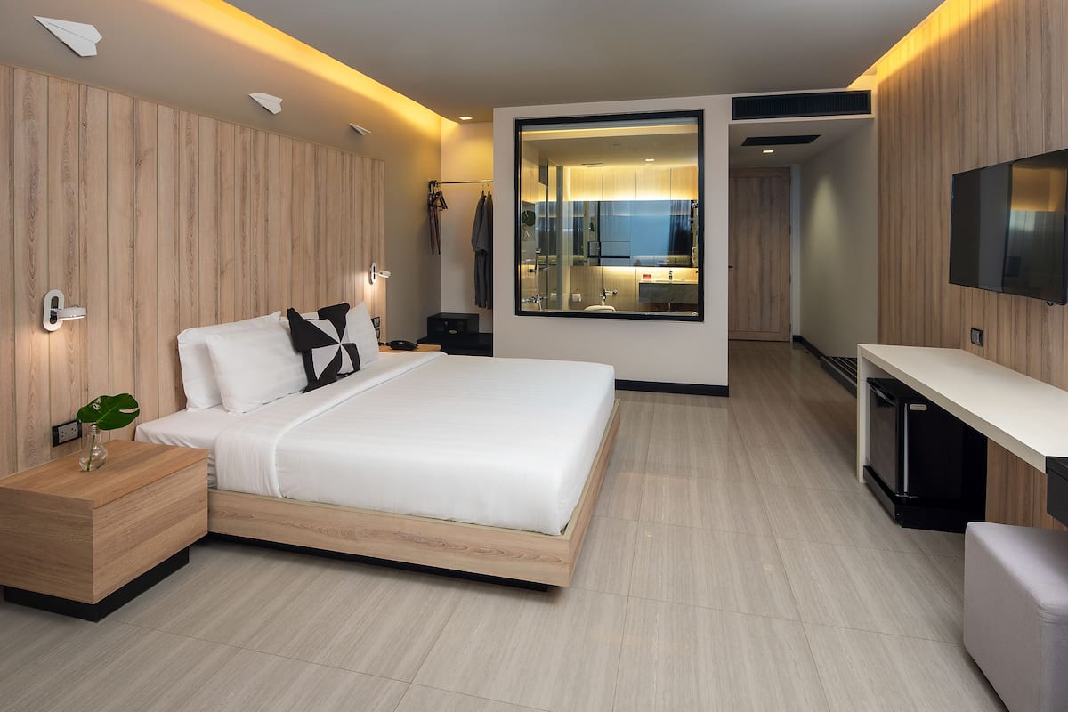 Une chambre d’hôtel moderne avec un lit doté d’une tête de lit en bois, une table de chevet avec un vase, un grand miroir, un téléviseur fixé au mur et un petit bureau.
