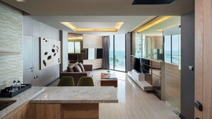 Une chambre d’hôtel moderne dotée d’un coin cuisine, d’un espace salon avec un canapé et des fauteuils, et d’une grande fenêtre offrant une vue sur l’océan.