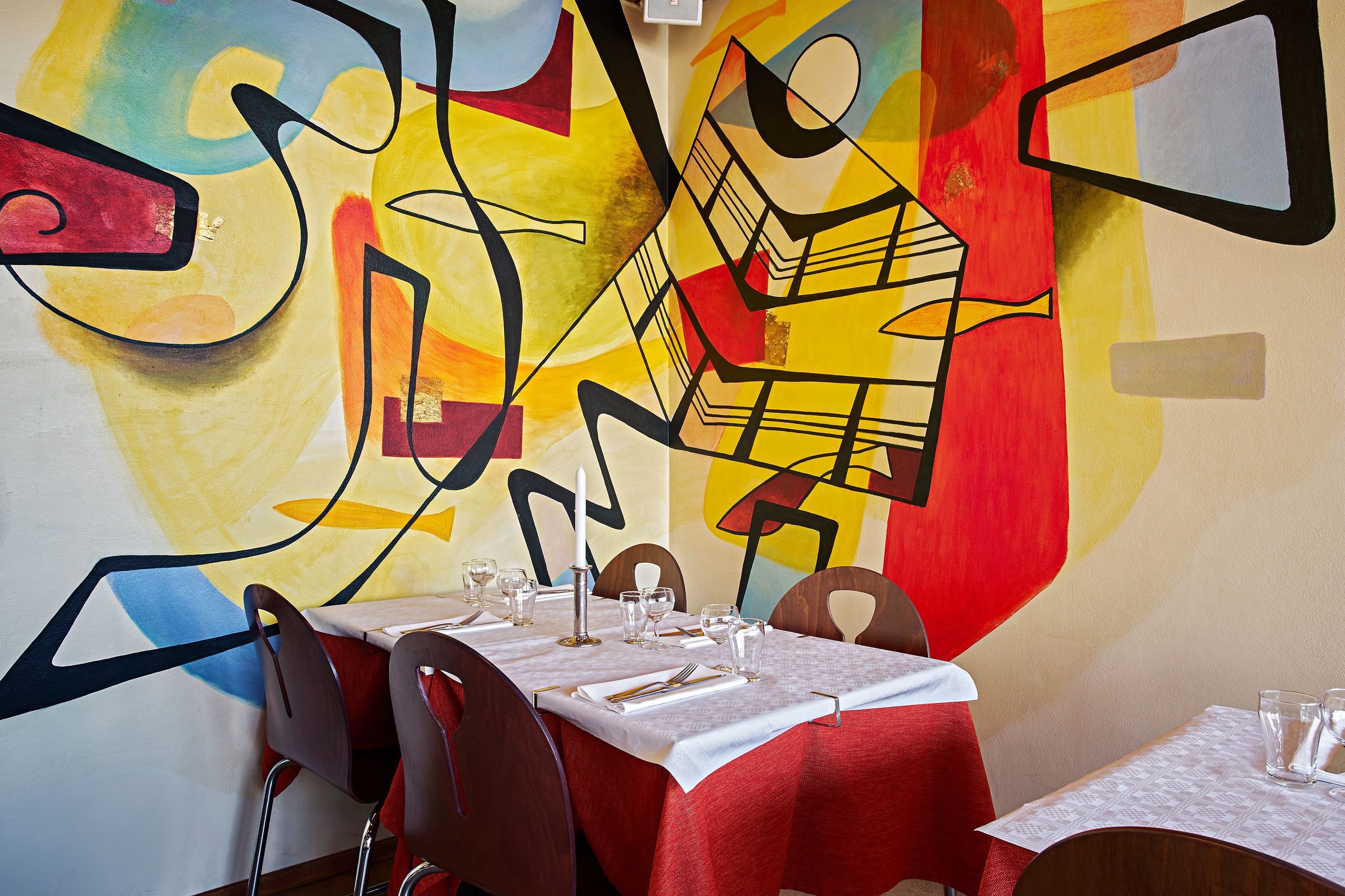A dining area with a table set for two, chairs, and a colorful abstract mural on the wall.