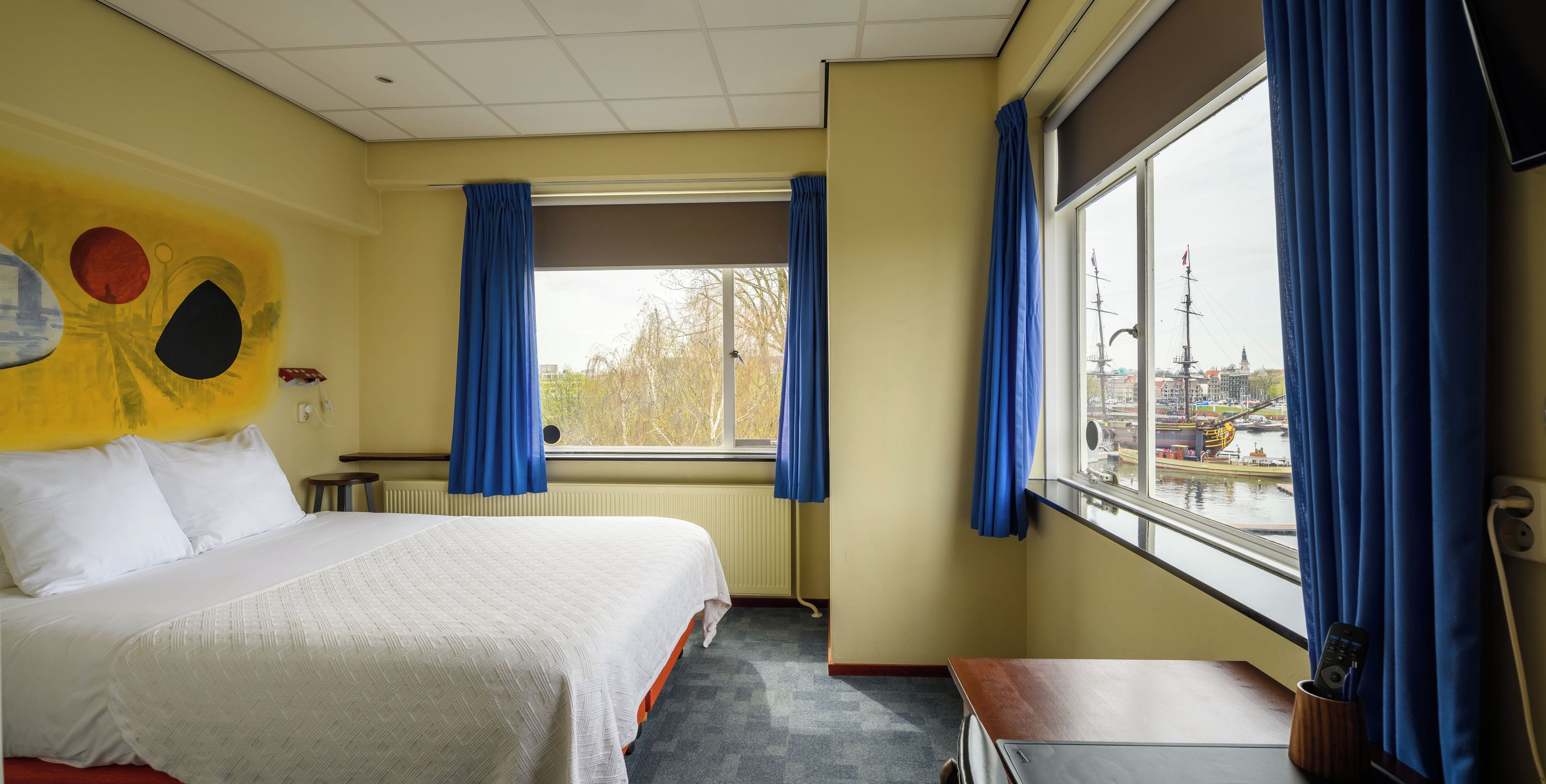Habitación de hotel con cama, ventana con cortinas y vista al puerto, además de un cuadro en la pared.