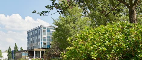 A modern building with a glass facade, surrounded by lush greenery and a body of water.