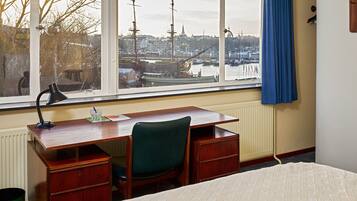 Een kamer met een houten bureau, een groene stoel, een bed en een raam met uitzicht op een haven en gebouwen.