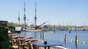 A waterfront restaurant with wicker furniture and a view of a tall ship docked at the harbor. - Pension Homeland (Amsterdam)