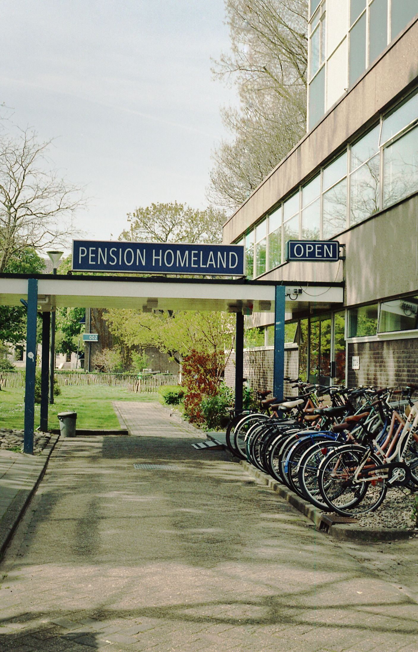 Entrada de um prédio com uma placa indicando 'PENSION HOMELAND' e uma área de estacionamento para bicicletas com várias bicicletas.