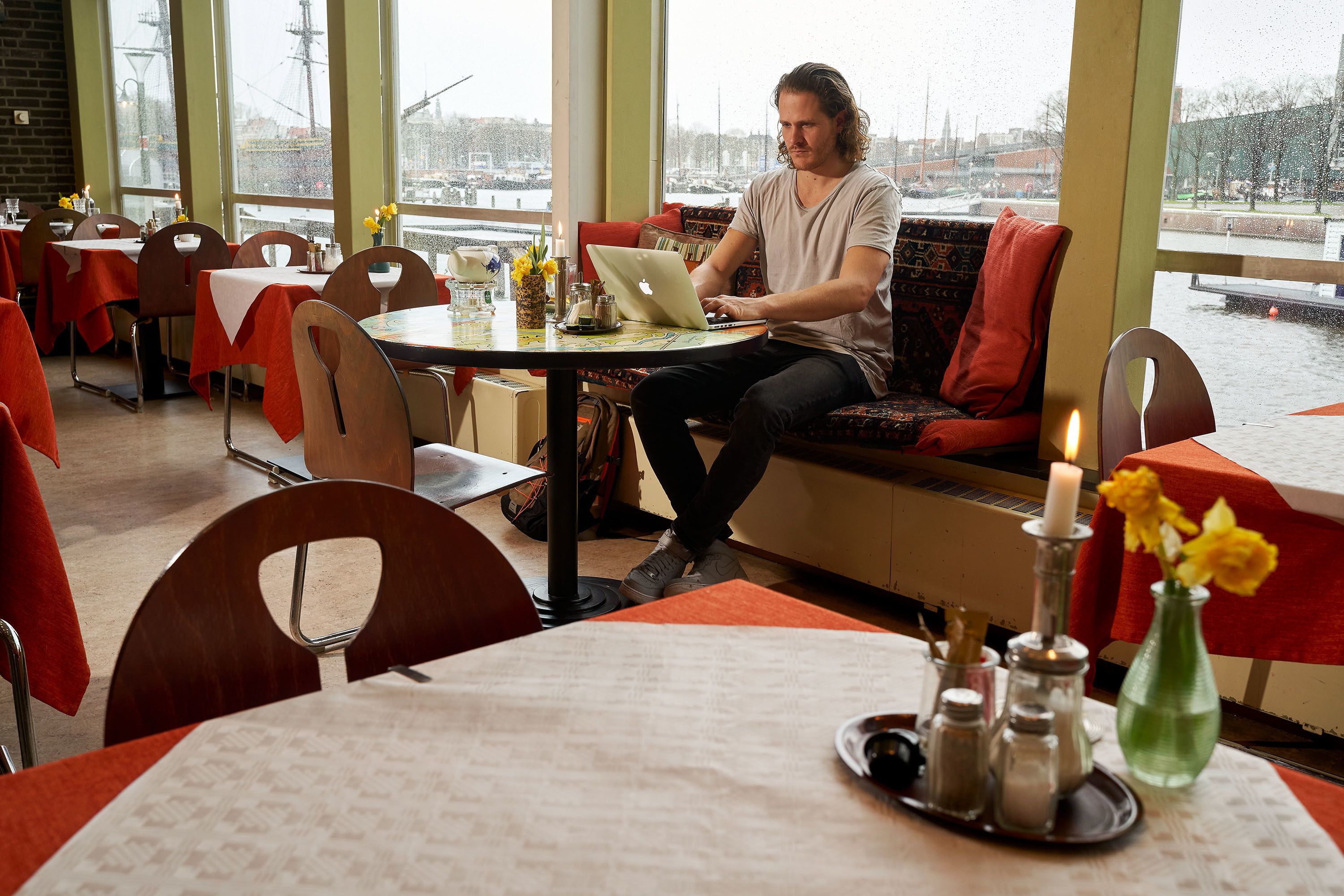 Un hombre trabajando en una computadora portátil en una mesa de restaurante con ventanas amplias, manteles rojos y vista a un cuerpo de agua.