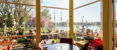 A restaurant with a view of a river and a ship, featuring round tables and chairs.