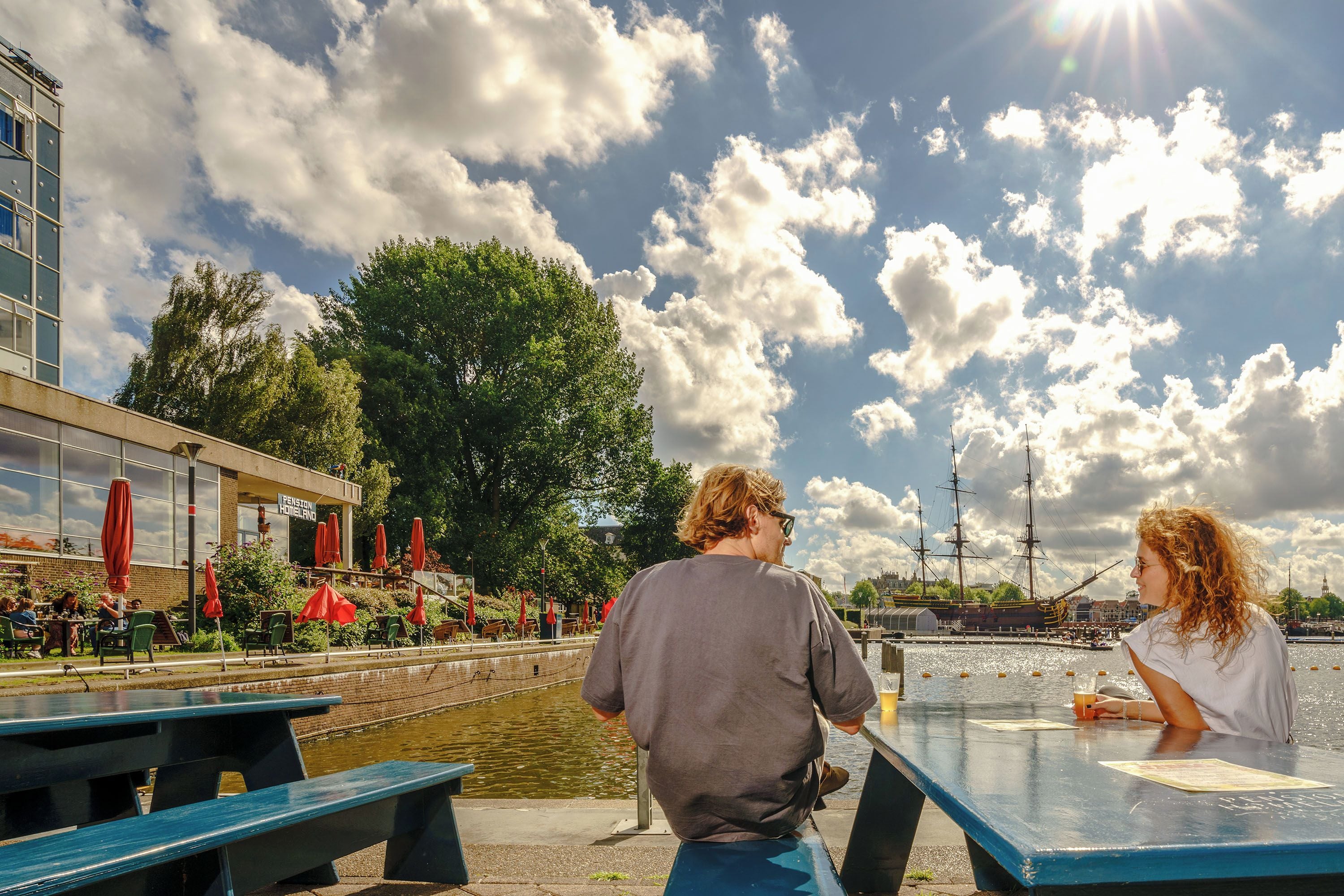 Een café aan de rivier met terras, uitzicht op het water en een historisch schip op de achtergrond.