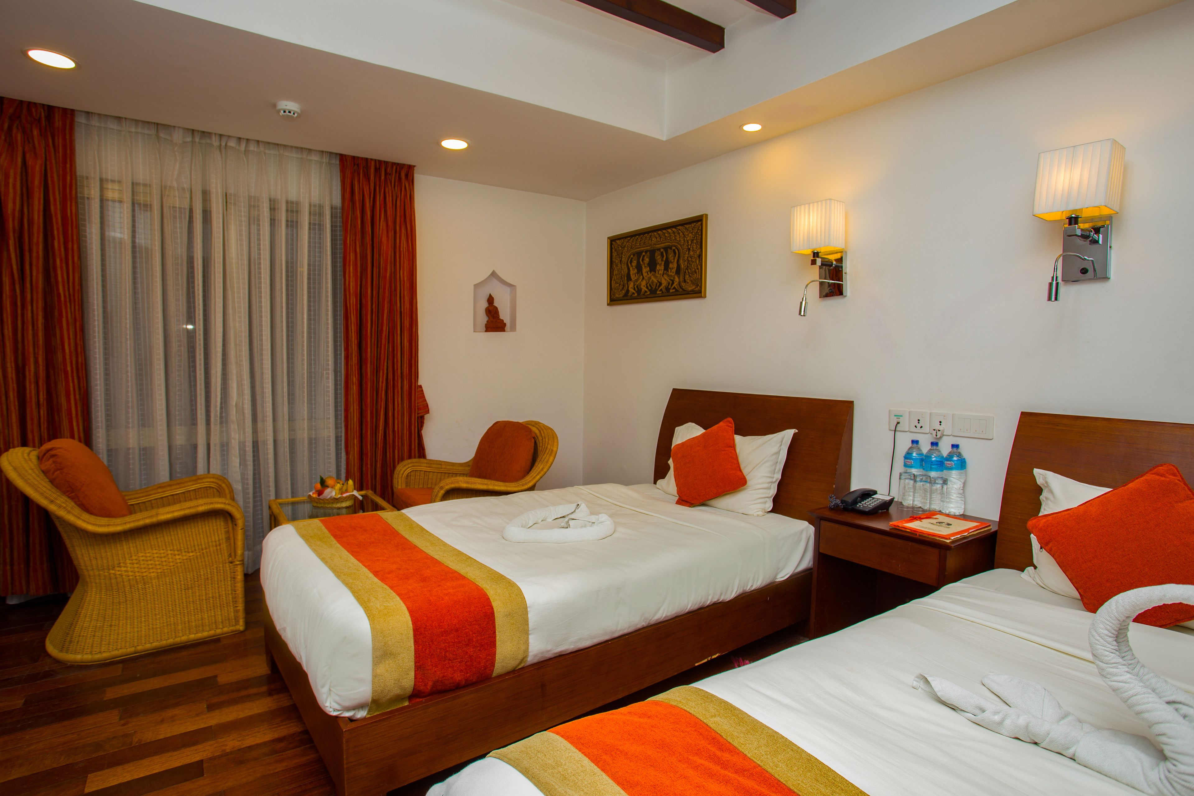 A hotel room with two beds, a wooden headboard, a nightstand, a chair, a window with curtains, and wall-mounted lamps.